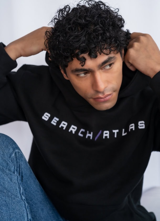 Young man in black SearchAtlas hoodie with curly hair, casual style, sitting indoors.