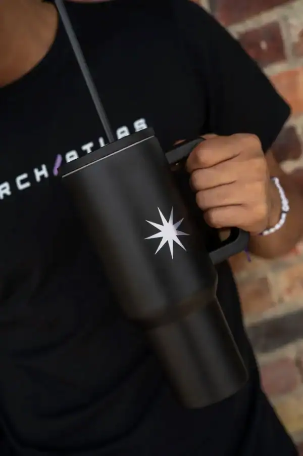 Black insulated travel mug with a star logo, held by a person wearing a black T-shirt with "SEARCH" lettering, against a brick wall background.